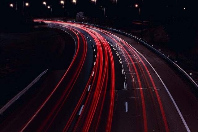 Fotografia noturna de longa exposição de uma rodovia. Em ambiente noturno, a rodovia de 3 pistas foi registrada sem nenhum carro aparecendo, porém vemos o vestígio que o trajeto de luz vermelha dos faróis deixou ao serem fotografados pela câmera no modo de longa exposição.