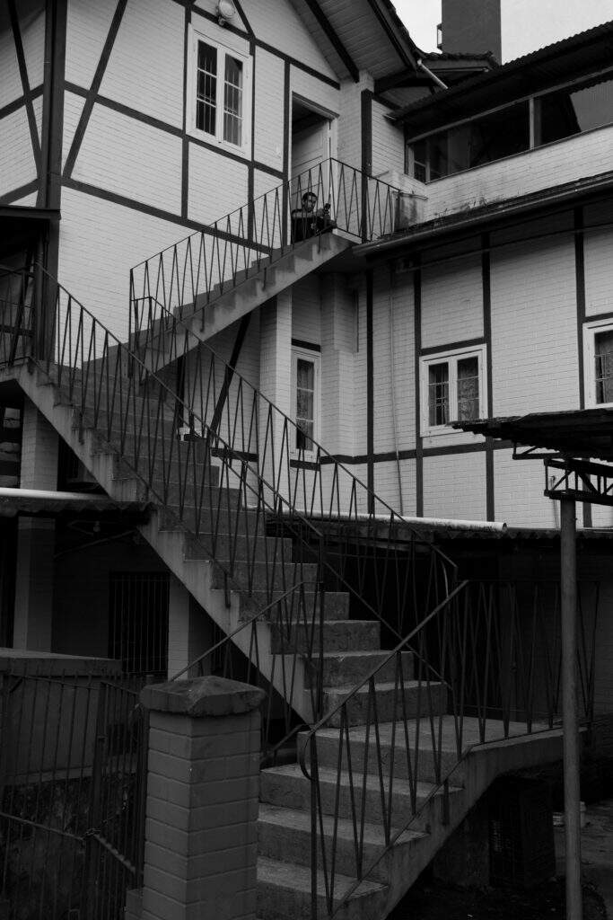 Audiodescrição da fotografia em preto e branco de uma construção imitação de enxainel com três andares. Três lances de escadas dão em uma porta, onde há um homem sentado.